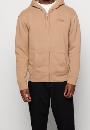Mann trägt beige Zip-up-Hoodie mit Fronttaschen und schwarze Hose vor einfachem Hintergrund.