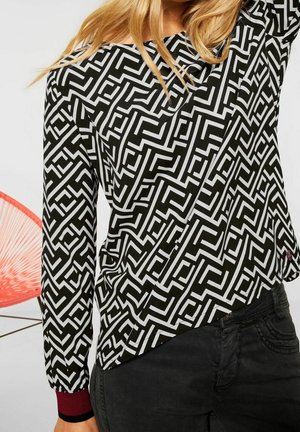 Nainen, jolla mustavalkoinen geometrinen kuvioitu pitkähihainen paita ja tummat farkut, vaaleat hiukset osittain näkyvissä, taustalla punainen tuoli.