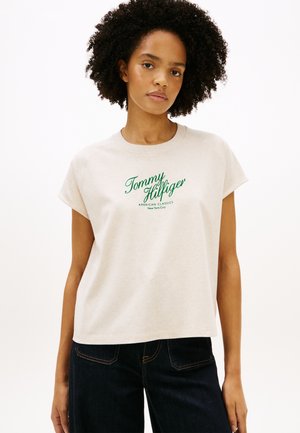 Frau mit lockigem Haar, die ein beiges Tommy Hilfiger T-Shirt mit grünem Text und dunkelblaue Jeans trägt und vor einem einfarbigen Hintergrund steht.