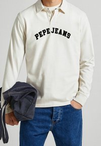 Mann in einem weißen Langarm-Polohemd mit dem Schriftzug "PEPE JEANS", blauen Jeans und hält eine gefaltete dunkelblaue Jeansjacke in der linken Hand.