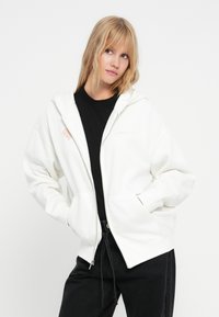 Hvit zip-up hettegenser med kengurulomme og ribbestrikkede mansjetter. Har brodert logo på brystet og er laget over en svart skjorte.