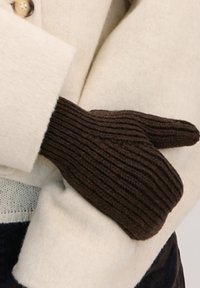 Brązowe dziergane mitenki z prążkowaną fakturą, noszone na beżowym wełnianym płaszczu. Mitenki są wyraźnie widoczne na tle jasnego materiału.