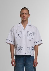Korte mouwen witte shirt met blauwe geborduurde accenten, twee borstzakken, bloempatronen en een relaxte pasvorm.