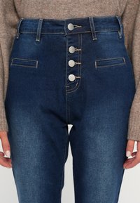 Jeans de tiro alto azul oscuro con cinco botones plateados, dos bolsillos frontales y un ligero desvanecimiento en los muslos. Hechos de material de mezclilla.