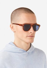 Sluneční brýle s želvovitou obrubou a tmavými skly, kulatého tvaru s tenkými nožičkami. Nosit se světle modrou mikinou s kapucí.