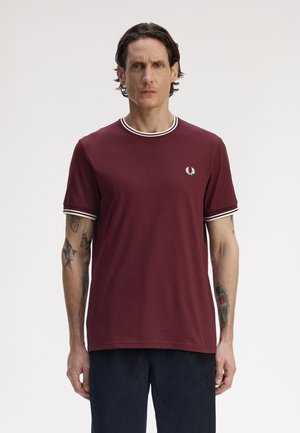 Homme tatoué portant un t-shirt bordeaux à manches courtes avec des rayures blanches sur les manches et le col, debout devant un fond uni.