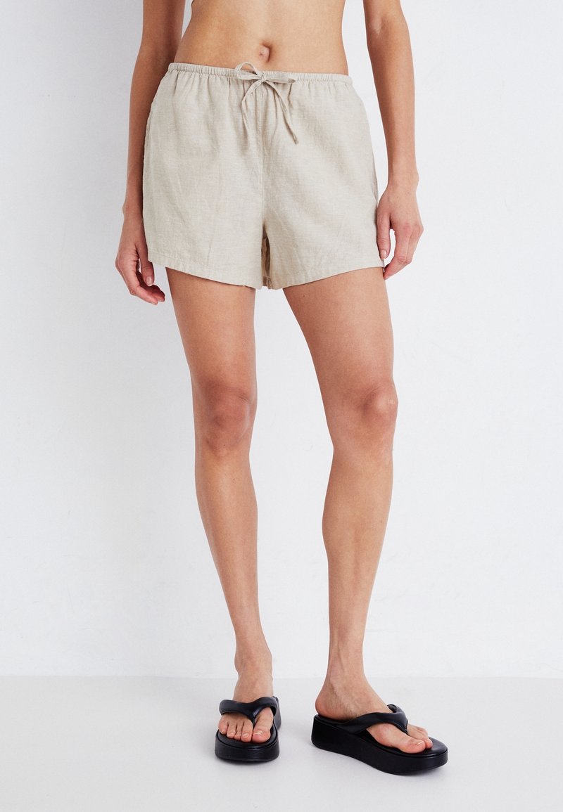 Personne portant un short beige à cordon et des tongs plateformes noires, debout devant un fond blanc.