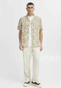 Chemise beige à manches courtes avec un motif abstrait marron clair, assortie à un pantalon en tissu léger et des baskets noires avec des semelles crème.