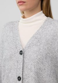 Grauer gestrickter Cardigan mit strukturiertem Muster, V-Ausschnitt und schwarzen Knöpfen, über einem hellen Rollkragenpullover getragen.
