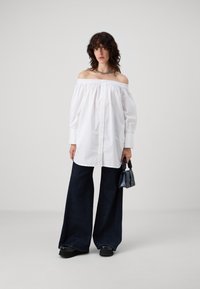 Blusa bianca off-the-shoulder con maniche lunghe e abbottonatura frontale, abbinata a jeans in denim scuro a gamba larga e una piccola borsa nera.