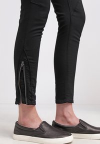 Černé skinny džíny se zipy u kotníků, které mají těsný střih a hladkou texturu. Kombinované s tmavými slip-on botami.