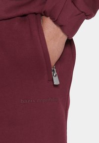 Bordeauxfarvede sweatpants har en lynlåslomme med et sølvfarvet træk og et diskret præget logo på stoffet nær lommen.