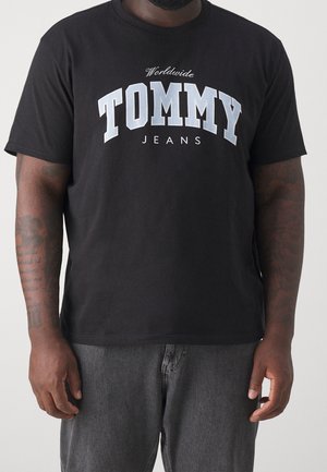 T-shirt noir à manches courtes avec le texte "Worldwide TOMMY JEANS" porté par une personne avec des bras tatoués et un jean en jean gris foncé.