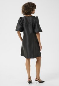 Svart satin klänning med puffärmar och en nyckelhålsdetalj i ryggen, med en slät yta och knälång fåll. Slingback-klackar bärs.