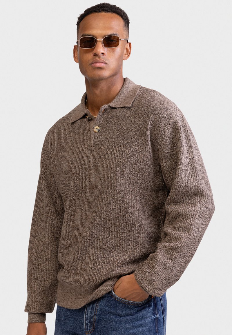 Homme portant un pull polo en maille marron à manches longues et des lunettes de soleil, avec une main dans la poche d'un jean bleu, devant un fond clair uni.