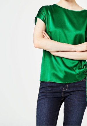 Femme portant un chemisier vert brillant à manches courtes et un jean bleu foncé, bras croisés, sur un fond blanc uni.