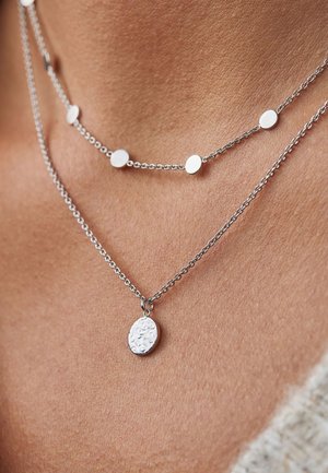Colliers en argent avec un pendentif ovale texturé et de petits détails en disque rond. Les chaînes sont délicates, réfléchissant la lumière avec une finition polie.