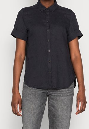 Personne portant une chemise noire à manches courtes avec col et boutons, et un jean gris clair, debout devant un fond clair uni.