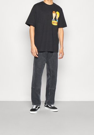 Camiseta de algodón negra con diseño gráfico que presenta dos personajes, combinada con unos jeans grises de corte holgado y zapatillas negras con suelas blancas.