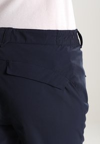 Close-up of navy blue pants with elastic waistband and flap pocket worn with light pink top.