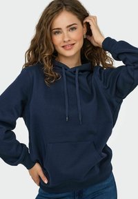 Hoodie bleu marine en matière douce avec une poche kangourou, capuche ajustable avec cordon de serrage et poignets côtelés. Le modèle pose avec une main dans la poche.