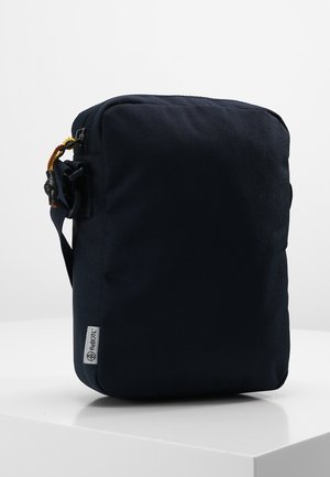 Kompaktowa ciemnogranatowa torba na ramię z regulowanym paskiem i małą metką z logo w dolnym lewym rogu, umieszczona na białej powierzchni.