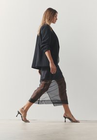 Blazer nero con maniche arrotolate, gonna nera trasparente con fodera marrone e sandali neri con tacco e cinturini, con un dettaglio decorativo.