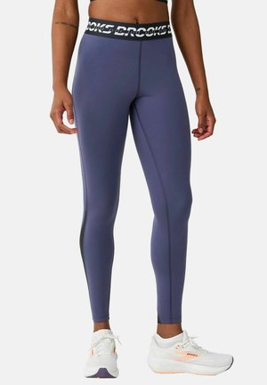 Ben iført blå atletiske leggings med sort og hvidt mærket taljebånd og hvide løbesko med orange accenter på en ensfarvet baggrund.