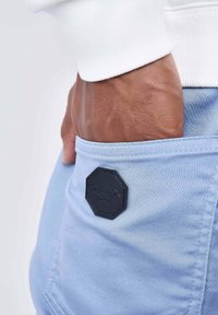 Mano con tonalità della pelle scura inserita nella tasca posteriore di pantaloni azzurri chiari con un patch logo ottagonale nero.