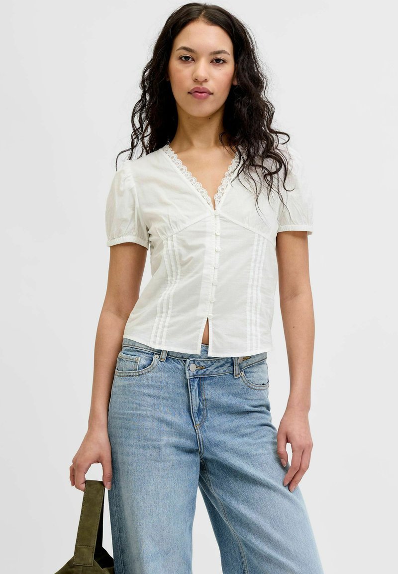 Jeune femme aux longs cheveux bouclés portant un chemisier blanc à manches courtes et un jean bleu clair, tenant un sac à main vert olive.