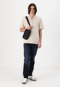 Beige polo shirt med krage, parret med mørkeblå jeans og hvide sneakers med sorte og grå accenter; bærende en lille sort crossbody taske.