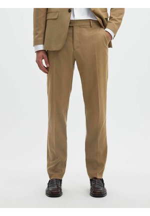 Homme portant un pantalon de costume beige et une veste assortie avec une chemise blanche, des chaussures de ville marron foncé, une main dans la poche, debout sur fond blanc.