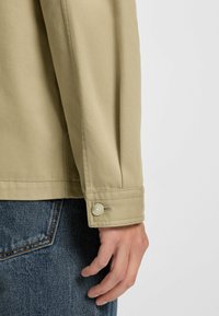 Khaki button-up jacket with long sleeves and a cuff detail, worn over blue denim. The fabric has a smooth texture.