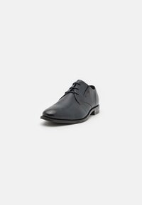 Chaussure noire en cuir avec une tige texturée, un design à lacets, un bout arrondi et un petit talon, présentant des coutures discrètes et une esthétique minimaliste.