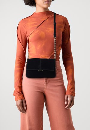 Černá semišová crossbody kabelka s klopou, řetězovým popruhem a hladkým povrchem. Modelka má na sobě hořčicově oranžové dlouhé tričko a světlé kalhoty.