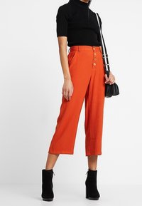 Roe rustkleurige cropped broeken met een voorkant knoopsluiting, gecombineerd met een zwart ribbeltrui met korte mouwen en zwarte enkellaarsjes.