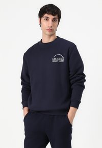 Marineblå sweatshirt med ribbet crew-halsudskæring og manchetter, prydet med et hvidt logo trykt på brystet. Stoffet ser blødt ud med en afslappet pasform.