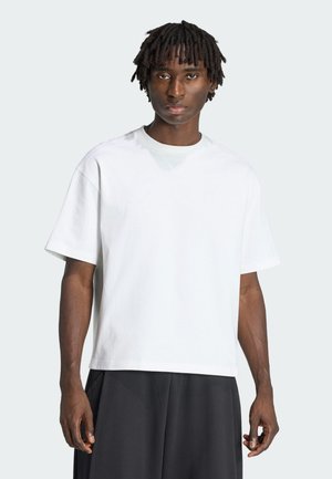 Junger Mann mit kurzen Dreadlocks, der ein einfaches weißes kurzärmeliges T-Shirt und lockere schwarze Hosen trägt, steht vor einem hellen Hintergrund.