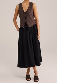 Brązowa lniana krótka kamizelka z dekoltem w szpic i wiązaniami, zestawiona z czarną spódnicą do kolan. Osoba ma na sobie brązowe buty na masywnym obcasie.