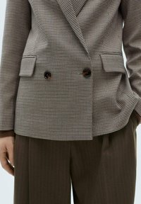 Blazer doppio petto a scacchi marroni con quattro bottoni, tasche laterali e polsini beige a contrasto. Abbinato a pantaloni marroni a righe sottili.