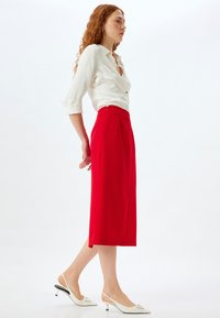 Roter Midirock mit strukturierter Silhouette, kombiniert mit einem weißen Hemd mit Knopfleiste. Das Outfit wird von eleganten, weißen High Heels ergänzt.