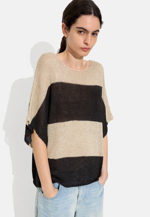 Młoda kobieta o ciemnych włosach związanych w kucyk, ubrana w luźny sweter w szerokie beżowo-czarne paski oraz jasnoniebieskie jeansy, z rękami w kieszeniach.