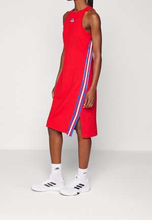 Femme portant une robe rouge sans manches avec des bandes latérales bleues, blanches et rouges, des chaussettes blanches Adidas et des baskets blanches Adidas, debout de profil.