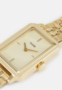 Montre rectangulaire en or avec un cadran crème texturé, des marqueurs minimalistes et un bracelet en maillons métalliques dorés. Elle possède une couronne sur le côté.