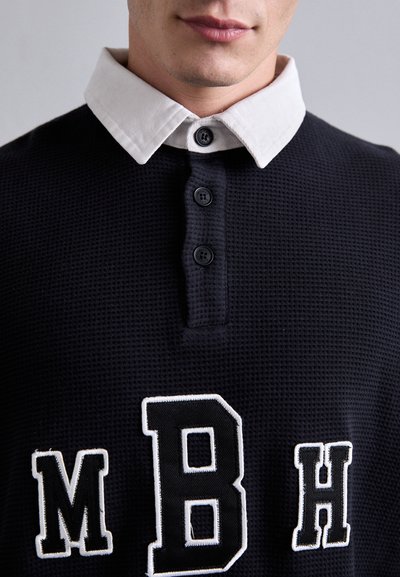 Marineblaues Poloshirt mit weißem Kragen, strukturiertem Stoff, drei Knöpfen und gestickten Buchstaben "MBH" auf der Vorderseite.