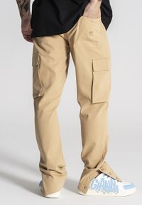 Beżowe spodnie cargo z wieloma kieszeniami, wykonane z trwałego materiału. Charakteryzują się luźnym krojem i zakończeniami w mankietach, w połączeniu z białymi tenisówkami.