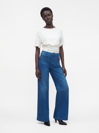 Femme debout portant un haut blanc à manches courtes rentré dans un jean bleu taille haute à jambes larges et des chaussures à talons noires à bout pointu.