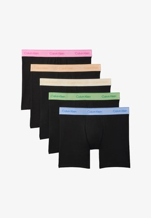 Oberkörperfreier Mann steht barfuß, trägt schwarze Boxershorts mit einem pinkfarbenen Calvin Klein Bund und richtet den Bund mit beiden Händen.