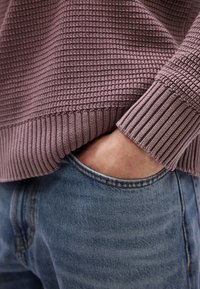 Άτομο που φοράει πουλόβερ με υφή σε μωβ απόχρωση, με μανίκια γυρισ μένα προς τα πάνω, το χέρι του είναι μέσα στην μπροστινή τσέπη ενός ανοιχτόχρωμου τζιν παντελονιού.