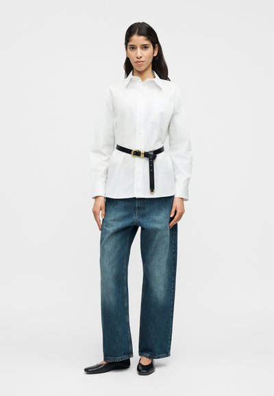 Λευκό πουκάμισο με κουμπιά συνδυάζεται με φαρδιά μπλε τζιν. Μια μαύρη ζώνη με χρυσή αγκράφα τονίζει τη μέση και μαύρα φλατ παπούτσια ολοκληρώνουν το ντύσιμο.